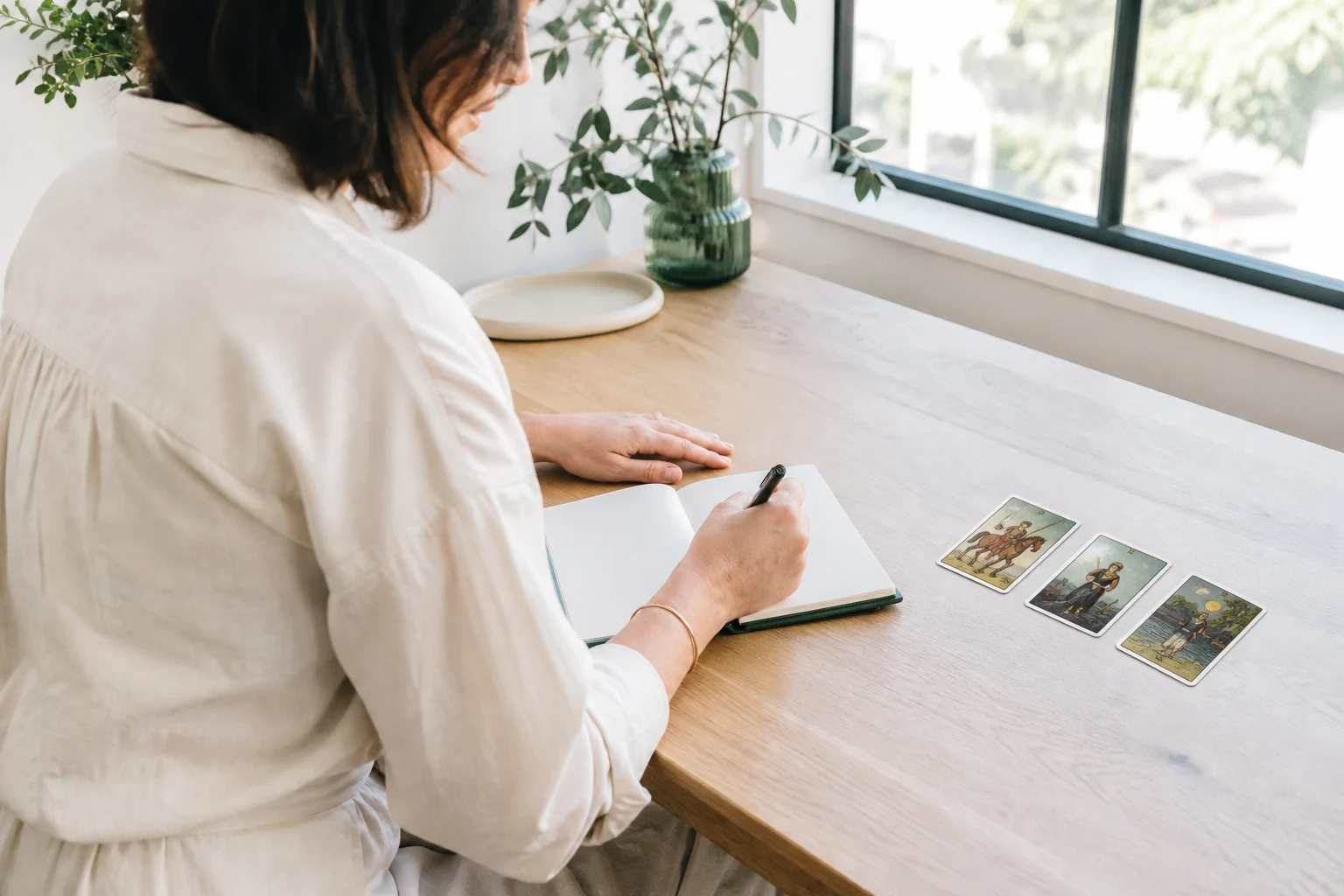 Consultante escribiendo una pregunta en un cuaderno junto a una tirada de tres cartas de tarot en una mesa luminosa